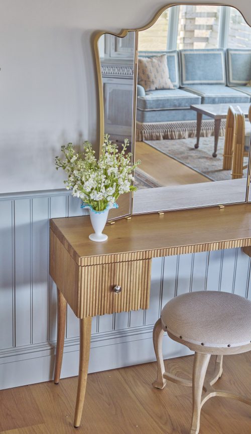 Homepage 10 Wooden vanity table with a round stool, featuring a vase of white flowers. The table has a mirror reflecting part of a bed. The setting has light-colored walls and paneling.