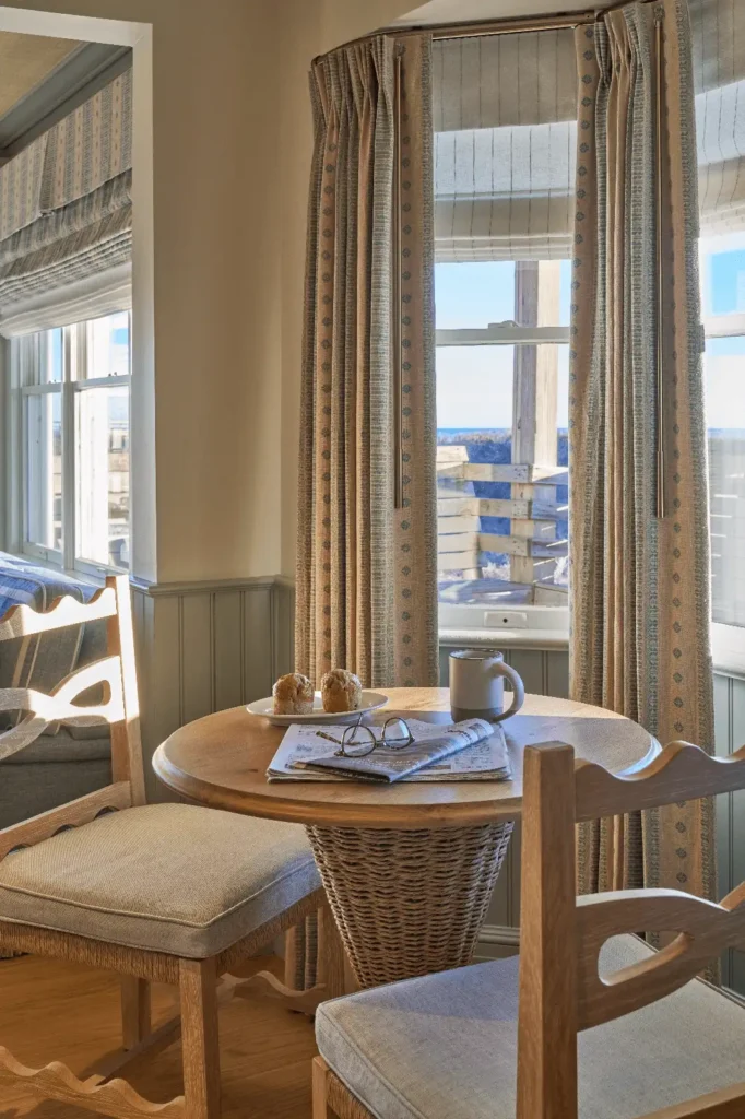 Charming corner of a room at The Sanderling Resort with a wooden table, two chairs, muffins, coffee, and a newspaper by the windows.